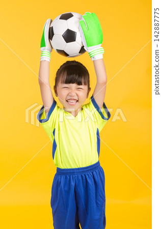The joyful East Asian girl in the soccer uniform cheers ecstatically, raising both hands clad in green goalkeeper gloves high above her head to celebrate with the black and white ball against the The joyful East Asian girl in the soccer uniform cheers ecstatically, raising both hands clad in green goalkeeper gloves high above her head to celebrate with the black and white ball against the 142887775