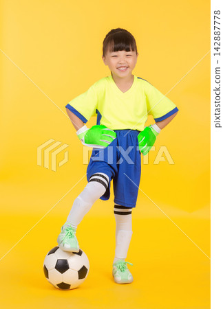 In a full-body action pose against the yellow studio background, the East Asian girl soccer player wearing green goalkeeper gloves rests her left foot casually on the soccer ball, looking confidently 142887778