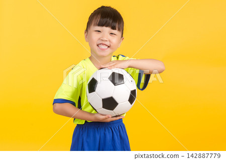 A bright-smiling East Asian elementary school girl stands confidently in front of a vibrant yellow background, a soccer ball tucked between her hands. A bright-smiling East Asian elementary school girl stands confidently in front of a vibrant yellow background, a soccer ball tucked between her hands. 142887779