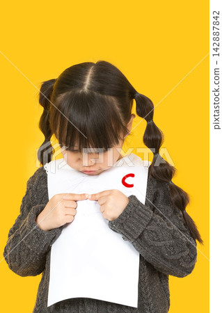 Little schoolgirl looking at her test paper with a bad grade C and feeling sad. Little schoolgirl looking at her test paper with a bad grade C and feeling sad. 142887842