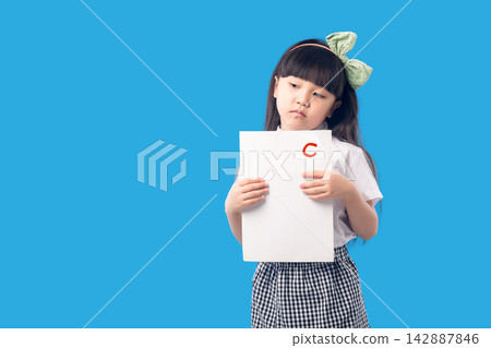 Sad little schoolgirl holding an exam paper with a low grade C on a blue background. Sad little schoolgirl holding an exam paper with a low grade C on a blue background. 142887846