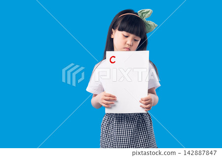 Sad little schoolgirl holding an exam paper with a low grade C on a blue background. Sad little schoolgirl holding an exam paper with a low grade C on a blue background. 142887847