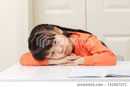 Bored and exhausted little girl leaning her head on the desk while looking at the camera. 142887871
