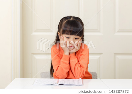 Unhappy little girl holding her chin in both hands and staring down at her notebook. Unhappy little girl holding her chin in both hands and staring down at her notebook. 142887872