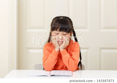 Unhappy little girl holding her chin in both hands and staring down at her notebook. Unhappy little girl holding her chin in both hands and staring down at her notebook. 142887873