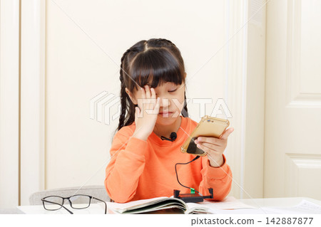 Stressed little East Asian girl holding her forehead while doing difficult homework at her desk. Stressed little East Asian girl holding her forehead while doing difficult homework at her desk. 142887877