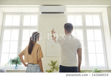 Couple using remote control to turn on air conditioner on wall of room at home 142888019