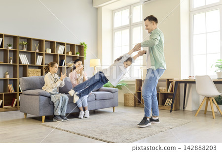 Father spinning daughter in cozy living room as mother and sister cheer happily Father spinning daughter in cozy living room as mother and sister cheer happily 142888203