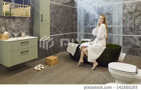 Beautiful young woman in white bathrobe sitting on edge of bathtub and preparing warm relaxing bath Beautiful young woman in white bathrobe sitting on edge of bathtub and preparing warm relaxing bath 142888205