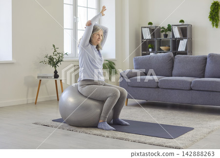 Healthy physical exercise, active workout of senior woman sitting on fitness ball in living room Healthy physical exercise, active workout of senior woman sitting on fitness ball in living room 142888263