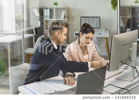 Business colleagues reviewing papers and data at desktop computer in glass office Business colleagues reviewing papers and data at desktop computer in glass office 142888338