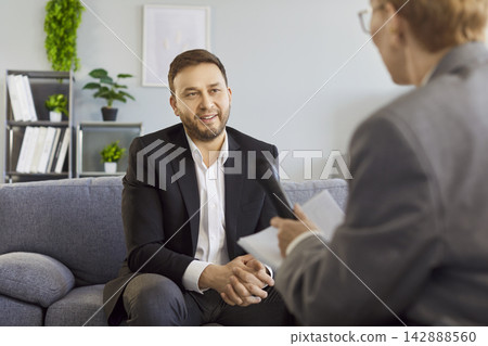 Businessman speaking in interview on sofa, interviewer with microphone in office Businessman speaking in interview on sofa, interviewer with microphone in office 142888560