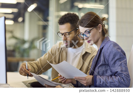 Focused coworkers analyzing papers together at desk in contemporary office Focused coworkers analyzing papers together at desk in contemporary office 142888930