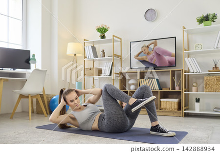 Young woman doing bicycle crunches at home, online workout training with TV coach Young woman doing bicycle crunches at home, online workout training with TV coach 142888934