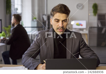 Serious businessman in suit working on laptop at modern open office workspace Serious businessman in suit working on laptop at modern open office workspace 142888936