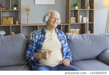 Content senior man listening to music at home on sofa with headphones comfortably Content senior man listening to music at home on sofa with headphones comfortably 142888939