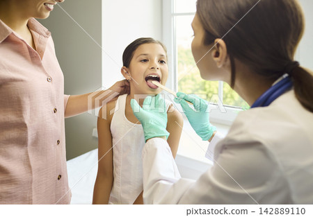 Pediatrician checks child throat with tongue depressor as mother nearby in clinic Pediatrician checks child throat with tongue depressor as mother nearby in clinic 142889110