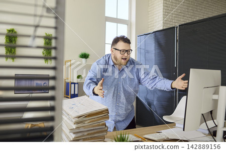 Stressed overweight office worker gesturing at computer with pile of paperwork 142889156
