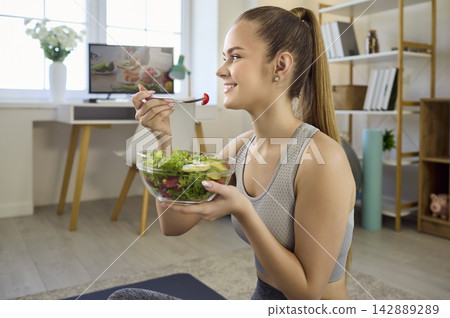 Fit woman eating salad at home after workout, healthy wellness lifestyle indoors Fit woman eating salad at home after workout, healthy wellness lifestyle indoors 142889289