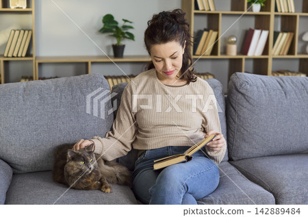 Calm woman reading a book beside her cat on sofa at home, relaxing cozy mood Calm woman reading a book beside her cat on sofa at home, relaxing cozy mood 142889484