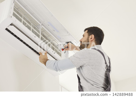 Skilled male mechanic or repairman repairing conditioner at client house on white wall Skilled male mechanic or repairman repairing conditioner at client house on white wall 142889488