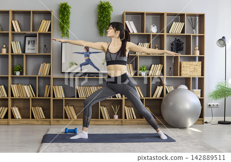 Female doing yoga warrior stretch in living room, home fitness via TV lesson Female doing yoga warrior stretch in living room, home fitness via TV lesson 142889511