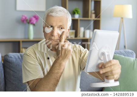 Senior man with under eye patches using mirror at home daily skincare routine 142889519