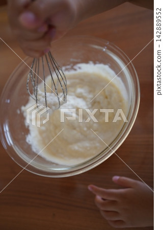 A child's hands mixing pastry batter in a glass bowl with a whisk. A child's hands mixing pastry batter in a glass bowl with a whisk. 142889552