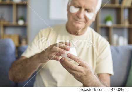 Senior man applying cream with under eye patches at home for skincare wellness Senior man applying cream with under eye patches at home for skincare wellness 142889603
