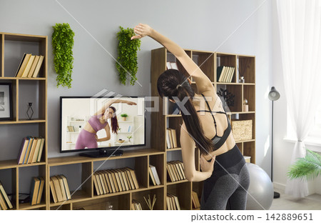 Young woman stretching at home with online fitness class on TV in cozy modern room Young woman stretching at home with online fitness class on TV in cozy modern room 142889651