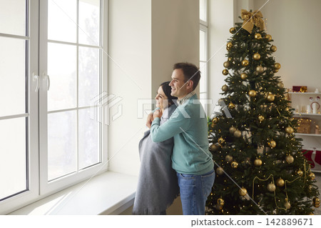On Christmas morning, couple is standing together by large window wrapped in plaid. On Christmas morning, couple is standing together by large window wrapped in plaid. 142889671