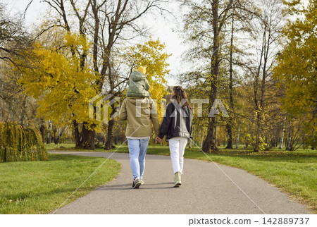Parents with daughter on shoulders strolling a winding autumn park pathway Parents with daughter on shoulders strolling a winding autumn park pathway 142889737