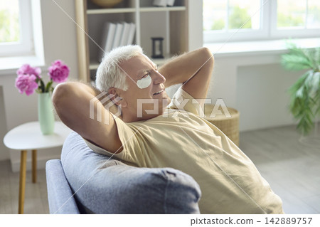 Senior man relaxing on sofa with under eye skincare patches at home for wellness Senior man relaxing on sofa with under eye skincare patches at home for wellness 142889757