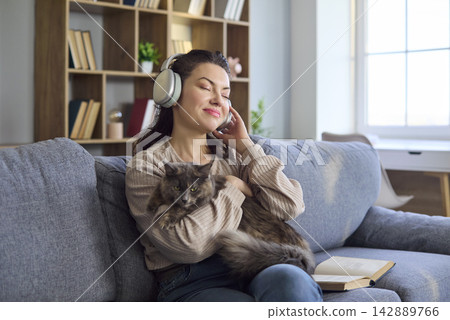 Happy woman in headphones enjoying music and cuddling cat on cozy home sofa Happy woman in headphones enjoying music and cuddling cat on cozy home sofa 142889766