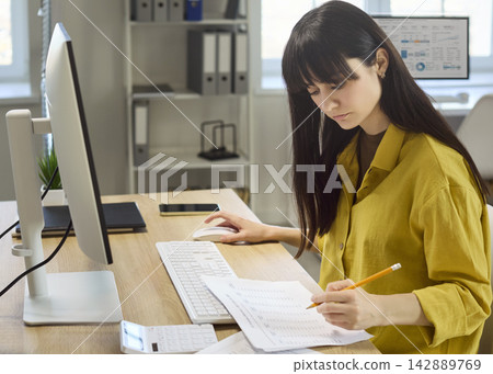Focused woman accountant reviewing budget paperwork at desktop computer in office Focused woman accountant reviewing budget paperwork at desktop computer in office 142889769