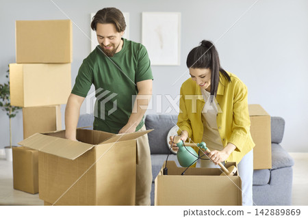 Smiling couple unpacking moving boxes in apartment, organizing home essentials 142889869