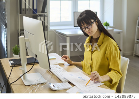 Focused woman accountant analyzing paperwork at office computer with calculator Focused woman accountant analyzing paperwork at office computer with calculator 142889873