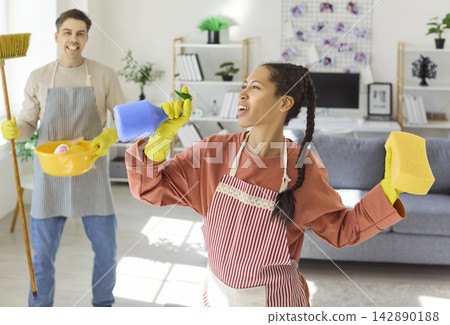 Playful couple cleaning living room, woman singing into spray bottle, fun day 142890188