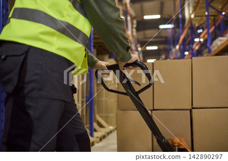 Closeup of warehouse worker steering pallet jack toward boxes on shelves Closeup of warehouse worker steering pallet jack toward boxes on shelves 142890297