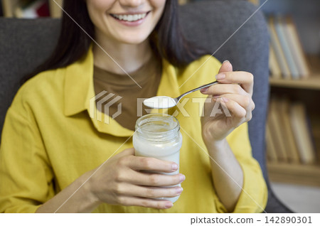Close up of woman eating yogurt with spoon at home, healthy snack concept Close up of woman eating yogurt with spoon at home, healthy snack concept 142890301