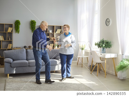 Happy senior couple dancing at home in bright living room, active retirement Happy senior couple dancing at home in bright living room, active retirement 142890335