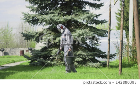 Person in protective coverall walking across suburban lawn while using handheld sprayer beside trees, with visible spray, daylight, safety focused outdoor maintenance scene Person in protective coverall walking across suburban lawn while using handheld sprayer beside trees, with visible spray, daylight, safety focused outdoor maintenance scene 142890395