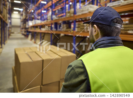 Warehouse worker transporting pallet boxes on pallet jack along industrial shelves 142890549