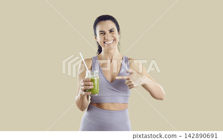 Fit woman in sportswear pointing at green smoothie on neutral studio background Fit woman in sportswear pointing at green smoothie on neutral studio background 142890691