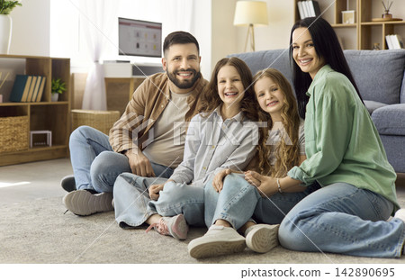 Parents with two daughters sitting on living room carpet, smiling at home Parents with two daughters sitting on living room carpet, smiling at home 142890695