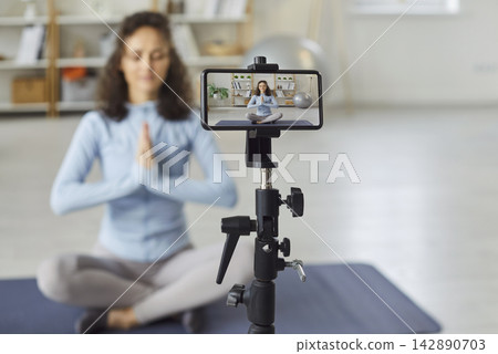 Sporty young woman recording video of online training in mobile phone camera doing yoga exercise. Sporty young woman recording video of online training in mobile phone camera doing yoga exercise. 142890703