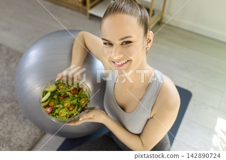 Smiling woman with fresh salad after home workout, promoting healthy lifestyle Smiling woman with fresh salad after home workout, promoting healthy lifestyle 142890724