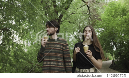 Man and woman enjoying ice cream cones in a sunny outdoor park, relaxed and casual mood, summer activities, eye-level perspective, green trees and concrete path. Man and woman enjoying ice cream cones in a sunny outdoor park, relaxed and casual mood, summer activities, eye-level perspective, green trees and concrete path. 142890725