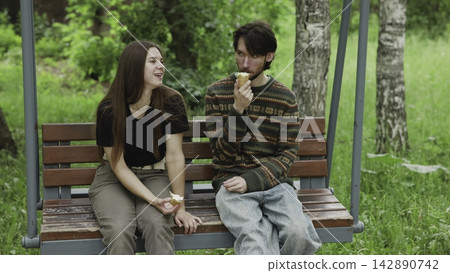 Young adults sharing ice cream on weathered wooden swing bench in leafy park under soft daylight, relaxed summer moment with casual clothing, greenery, and gentle smiles Young adults sharing ice cream on weathered wooden swing bench in leafy park under soft daylight, relaxed summer moment with casual clothing, greenery, and gentle smiles 142890742