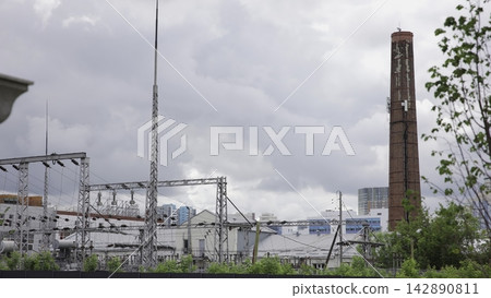 Industrial electrical substation beside weathered brick chimney under overcast sky, wide panorama showing power infrastructure, utility equipment, metal lattice towers, urban industry and muted Industrial electrical substation beside weathered brick chimney under overcast sky, wide panorama showing power infrastructure, utility equipment, metal lattice towers, urban industry and muted 142890811